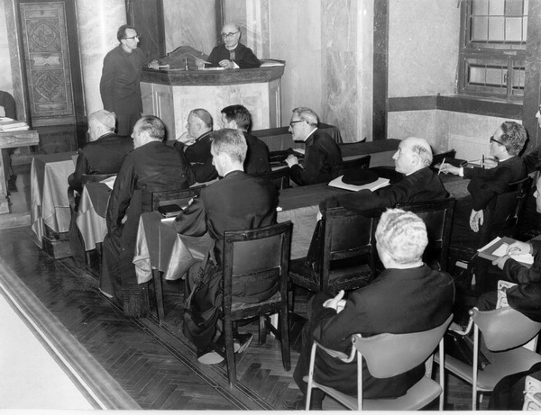 mons. giacomo biffi siede di fronte ai preti ripresi seduti in ascolto