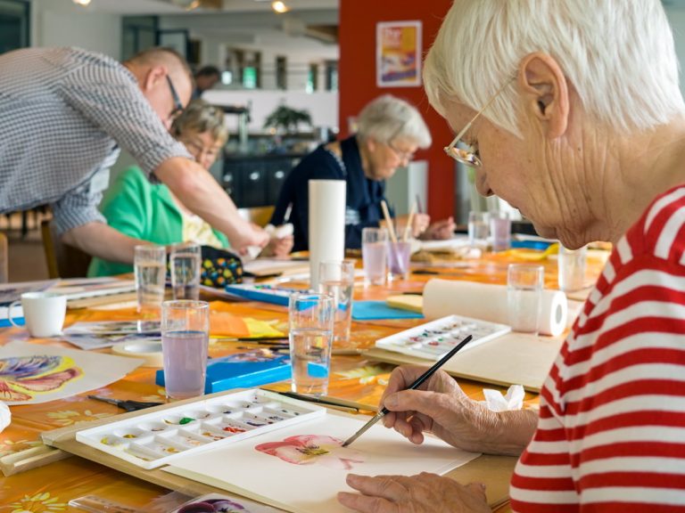 Woman in striped red and white shirt working on watercolor painting at table with other students in spacious studio.