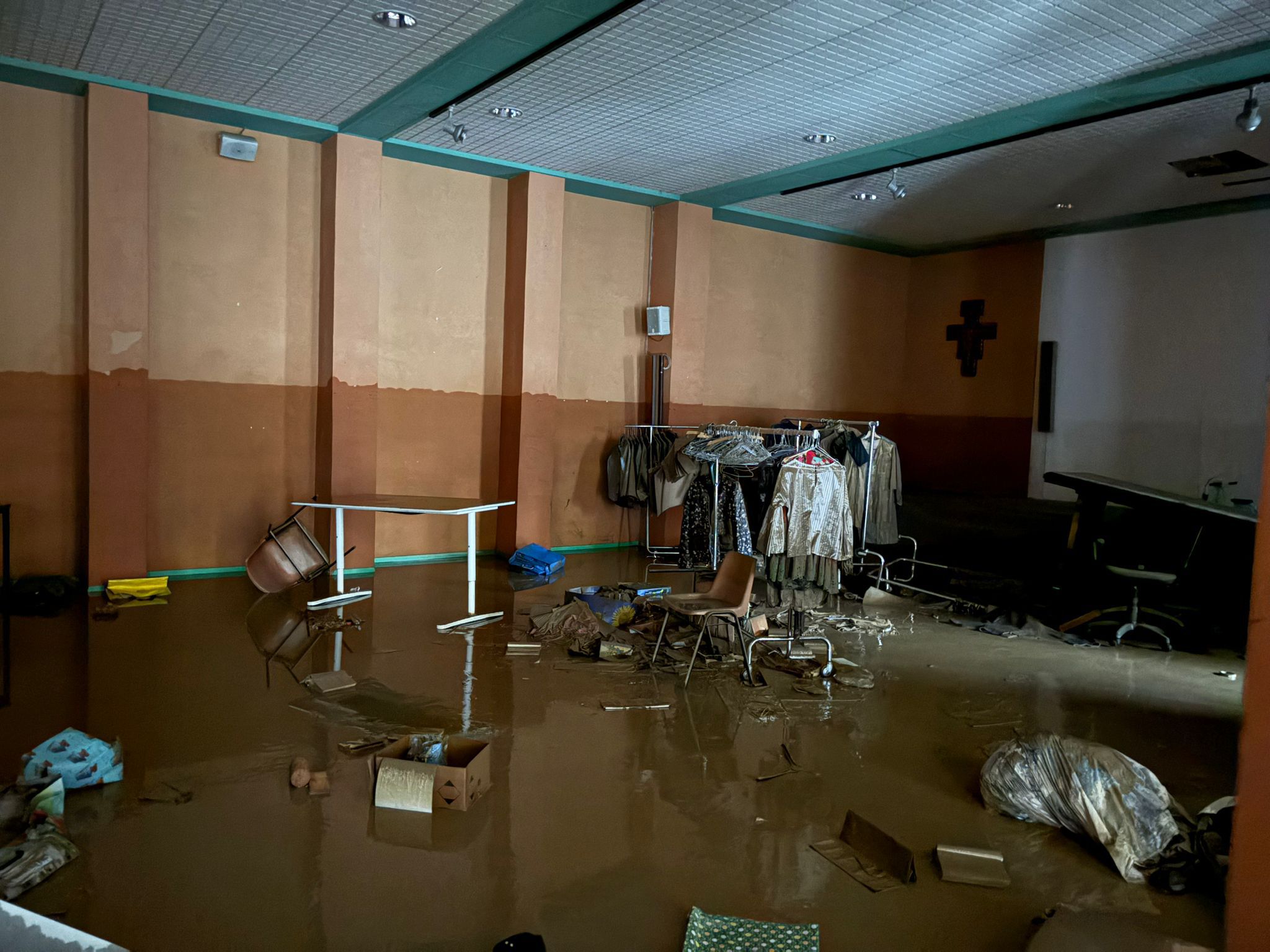 Un locale parrocchiale con guardaroba invaso dal fango, con sedie e arredi rovesciati e vestiti danneggiati dall’alluvione.