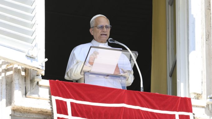 Papa Leone XIV in abito bianco parla da un leggio affacciato a una finestra, sopra un drappo rosso decorato con croci bianche.