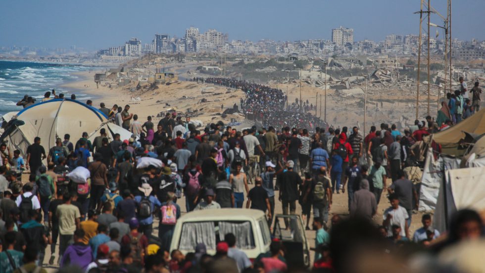 Una folla di persone cammina lungo una strada sabbiosa sulla costa, con tende e rovine sullo sfondo, in direzione della città in lontananza.