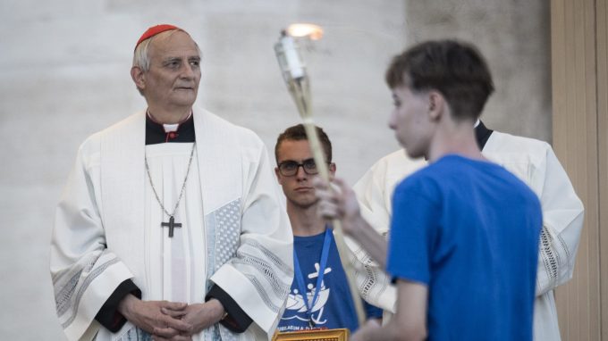 Il cardinale Matteo Zuppi in abiti liturgici osserva un giovane che regge una torcia accesa durante una cerimonia religiosa.