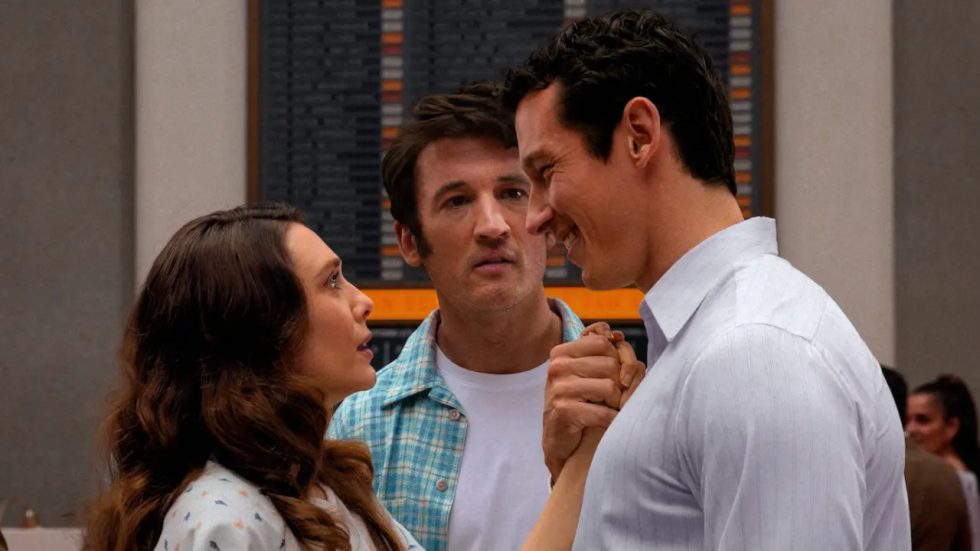 Una donna e un uomo si tengono le mani sorridendo in un ambiente interno, mentre un altro uomo dietro di loro osserva la scena con espressione preoccupata