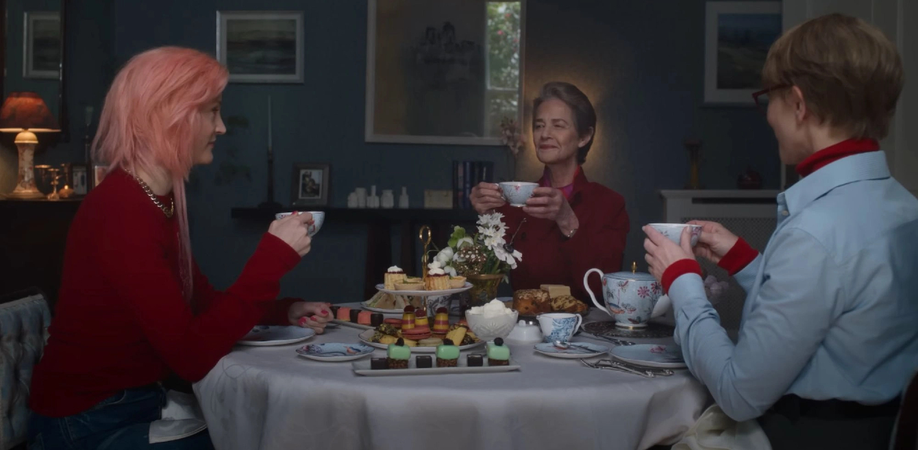 Tre donne sedute attorno a un tavolo apparecchiato per il tè, ciascuna con una tazza in mano; al centro dolci e stoviglie, in un’atmosfera raccolta e conviviale