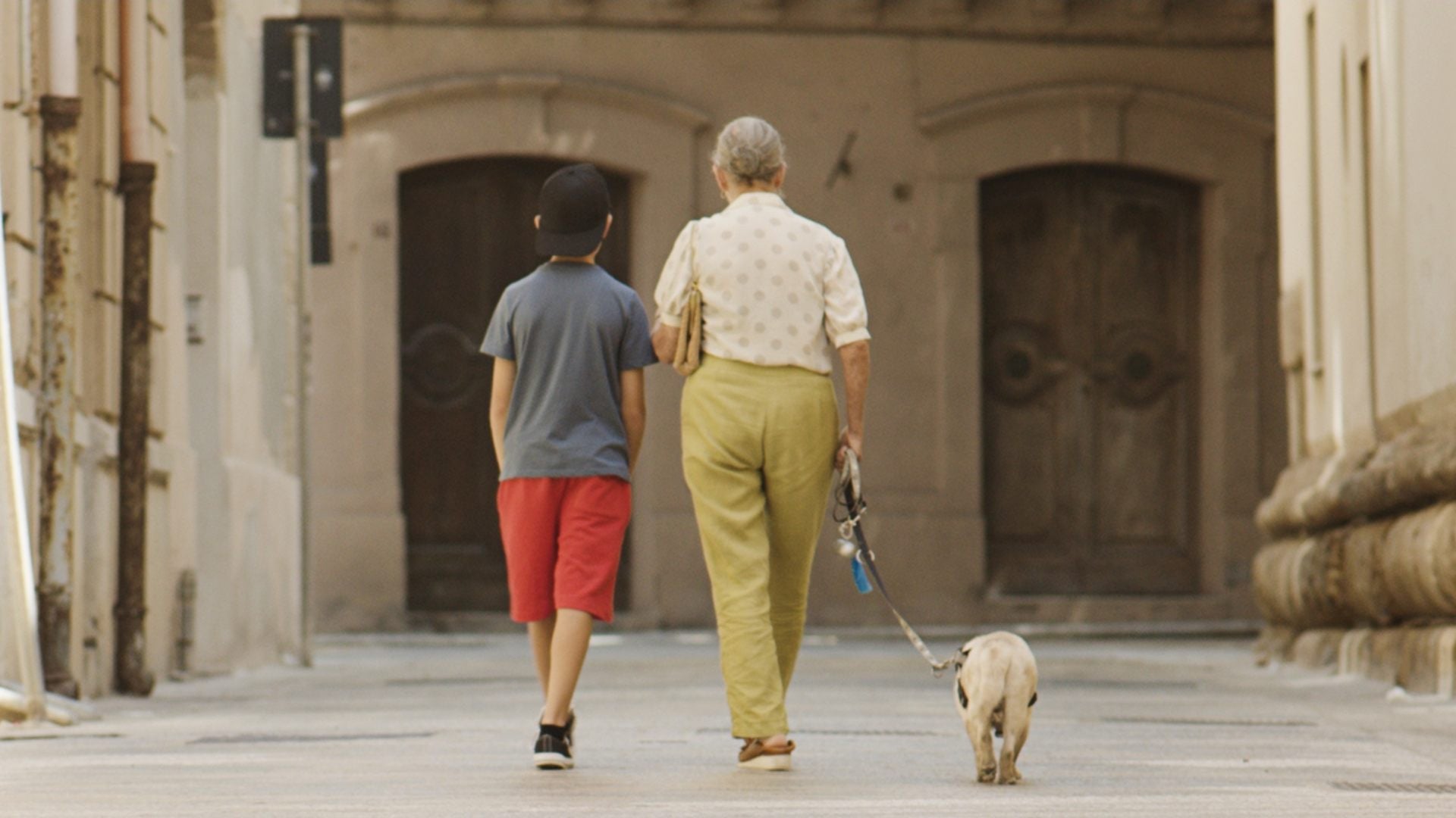 Un bambino e un’anziana donna camminano di spalle lungo una strada silenziosa tra edifici storici; il bambino indossa una maglietta scura e pantaloncini rossi, mentre la donna tiene al guinzaglio un piccolo cane, con grandi portoni in legno sullo sfondo.