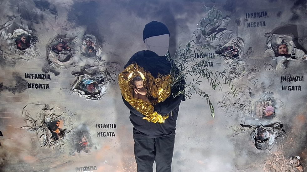 Installazione artistica che rappresenta una figura senza volto che regge un bambolotto avvolto in una coperta termica dorata e un ramo d’ulivo; sullo sfondo, un muro grigio simulato con fori da cui emergono fotografie di bambini, accompagnate dalla scritta ‘Infanzia negata’ ripetuta più volte