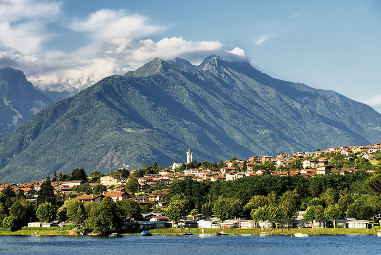Veduta di Colico sul Lago di Como con il paese affacciato sull’acqua, le case dai tetti rossi e la chiesa al centro, incorniciati dalle montagne verdi della Valchiavenna lungo il percorso del Cammino di San Colombano.
