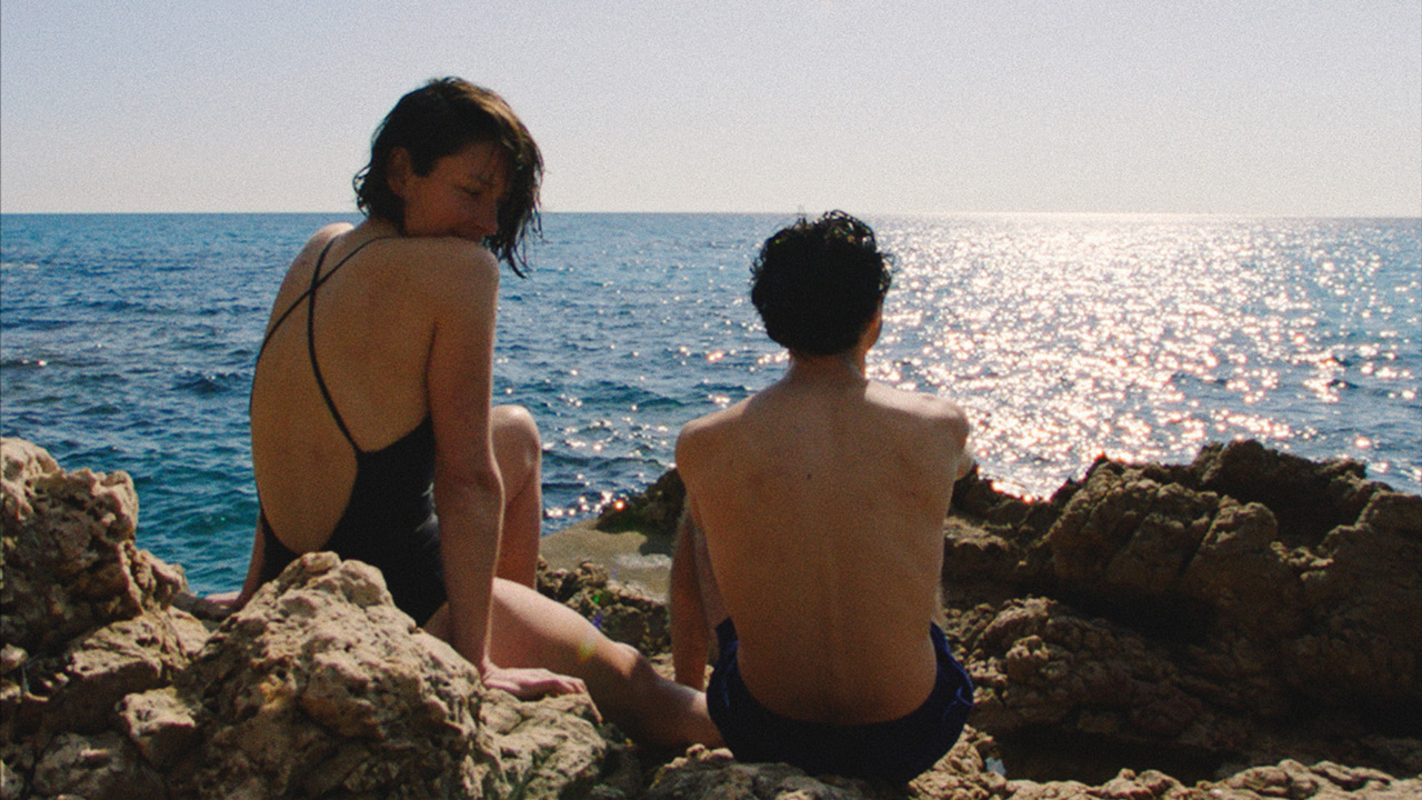 Scena del film ‘E.1027 – Eileen Gray e la casa sul mare’ con due personaggi seduti sugli scogli davanti al Mediterraneo vicino alla villa modernista E.1027