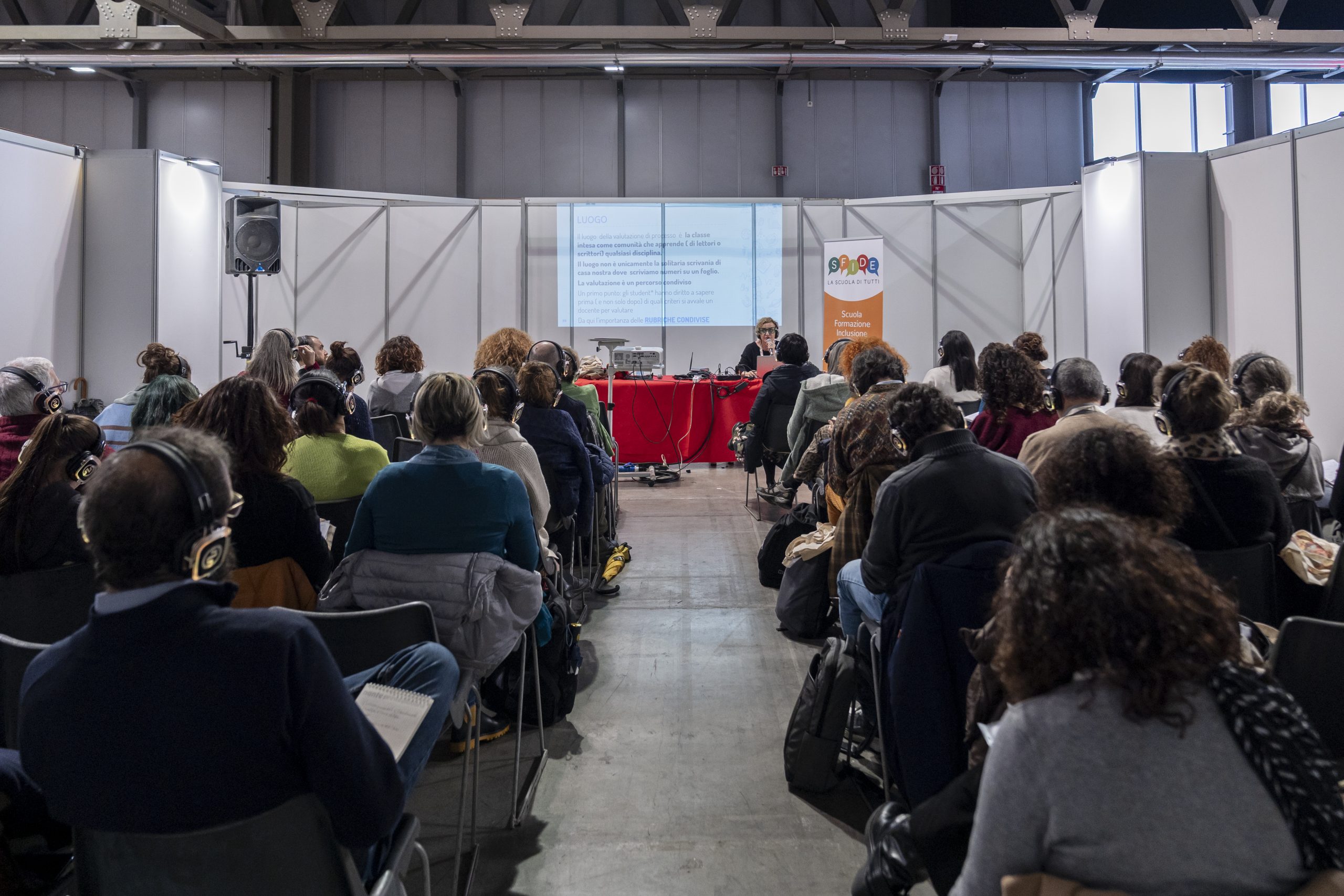 Pubblico seduto ascolta un incontro con cuffie per la traduzione simultanea durante un evento della fiera Fa’ la cosa giusta!, con una relatrice al tavolo e una presentazione proiettata sullo schermo.
