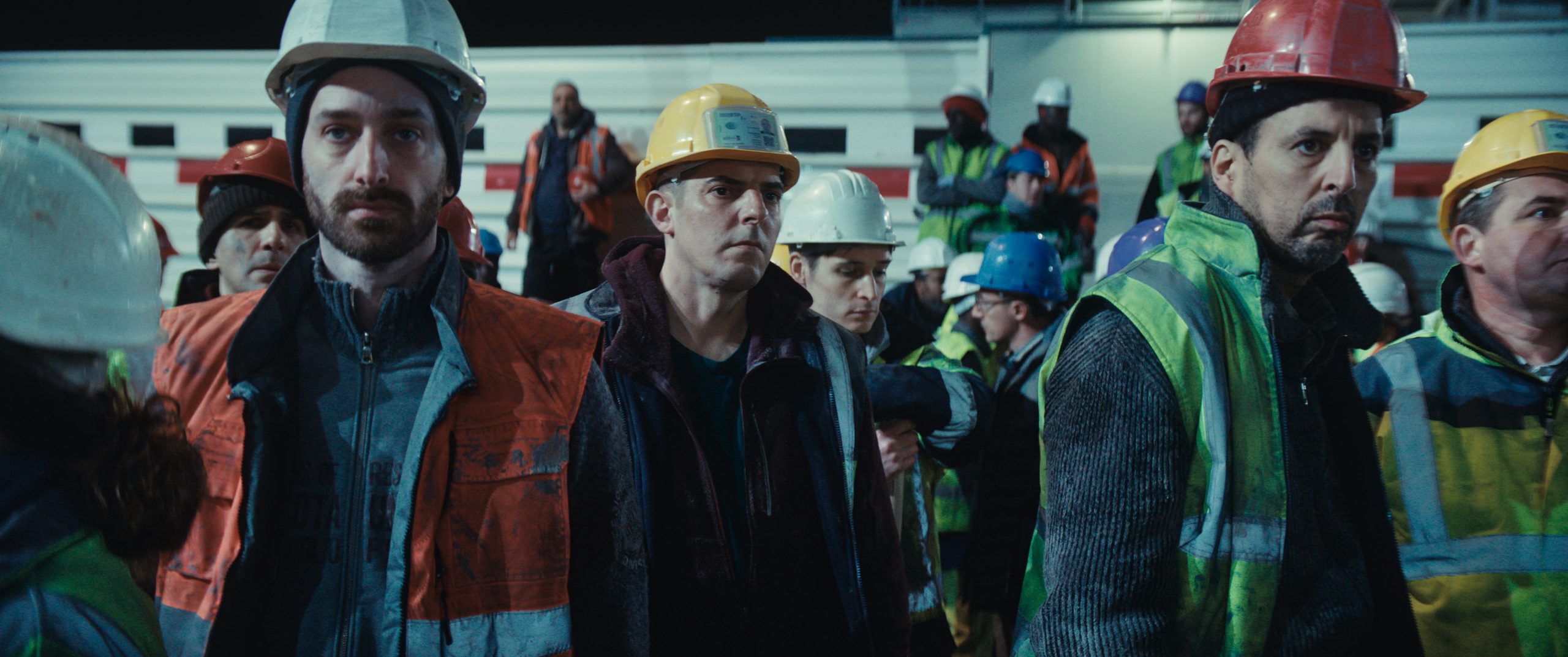 Gruppo di operai con caschi protettivi e giubbotti ad alta visibilità riuniti in un cantiere industriale in una scena del film Grand Ciel.