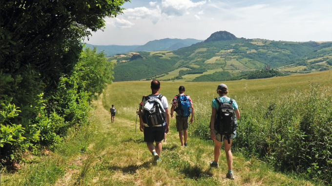 Escursionisti con zaini camminano lungo un sentiero erboso sulle colline della Val Trebbia, nel tratto del Cammino di San Colombano raccontato nel podcast, con campi coltivati e montagne sullo sfondo sotto un cielo estivo.