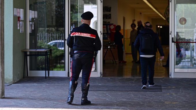 Fotografia di un Carabiniere in uniforme di spalle, con la scritta 'CARABINIERI' visibile sulla giacca, mentre cammina verso l'ingresso a vetri di un edificio. Un uomo con zaino lo precede ed entra nel corridoio di una scuola. Altre figure sfocate sono visibili all'interno
