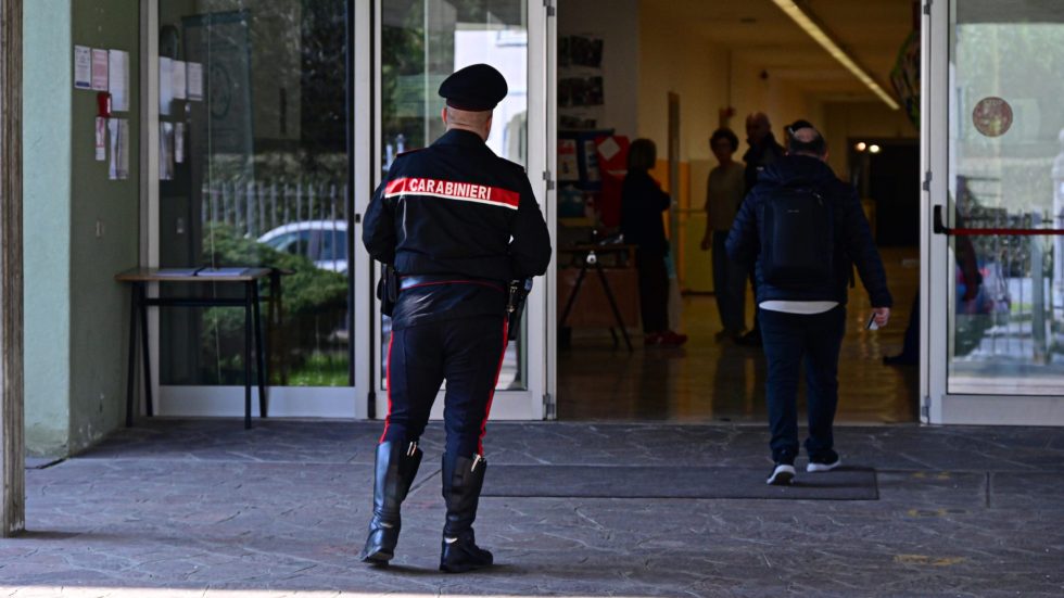 Fotografia di un Carabiniere in uniforme di spalle, con la scritta 'CARABINIERI' visibile sulla giacca, mentre cammina verso l'ingresso a vetri di un edificio. Un uomo con zaino lo precede ed entra nel corridoio di una scuola. Altre figure sfocate sono visibili all'interno