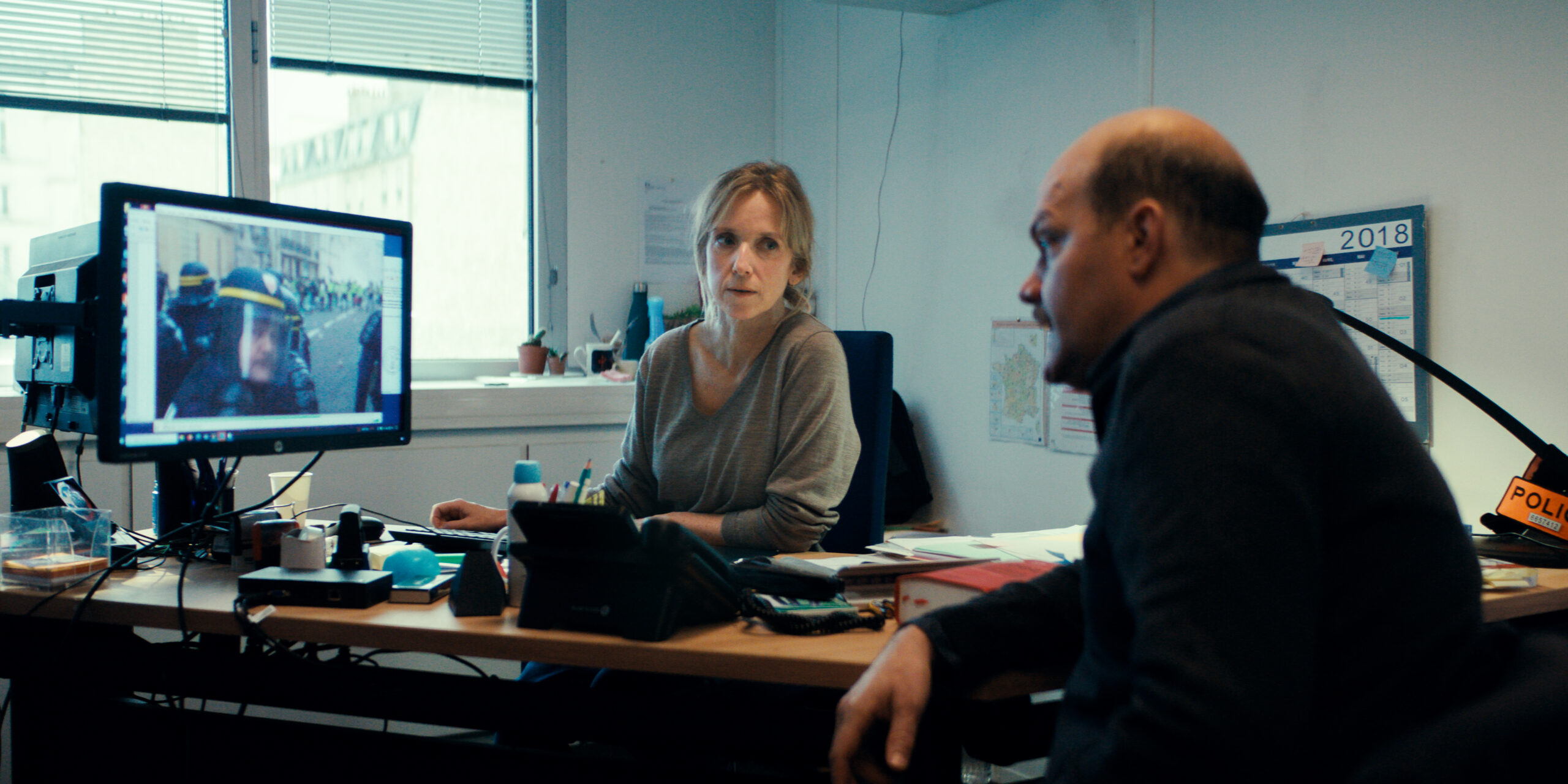 Una scena del film Il caso 137, dove due investigatori in ufficio osservano un monitor con immagini di scontri di piazza, tra scrivanie ingombre e atmosfera da indagine.