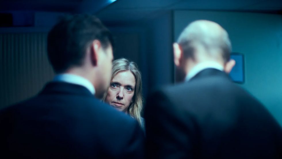 Una scena del film Il caso 137, dove un donna al centro di un corridoio osserva tra due uomini in abito scuro, scena dai toni freddi e atmosfera di tensione.