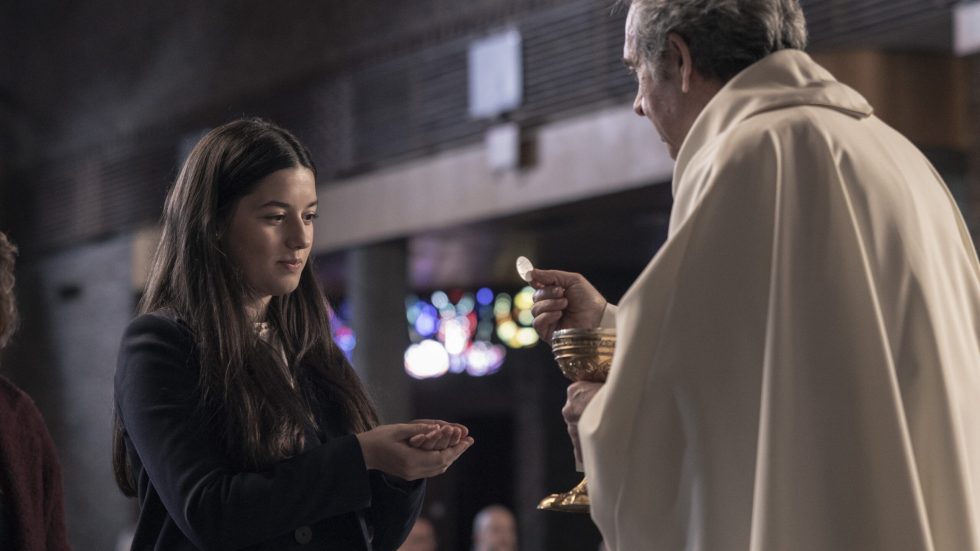 Primo piano di una ragazza che riceve l'ostia da un sacerdote durante una funzione religiosa in una chiesa. La ragazza tiene le mani giunte per ricevere la comunione mentre il sacerdote, di profilo, sorregge una pisside dorata.