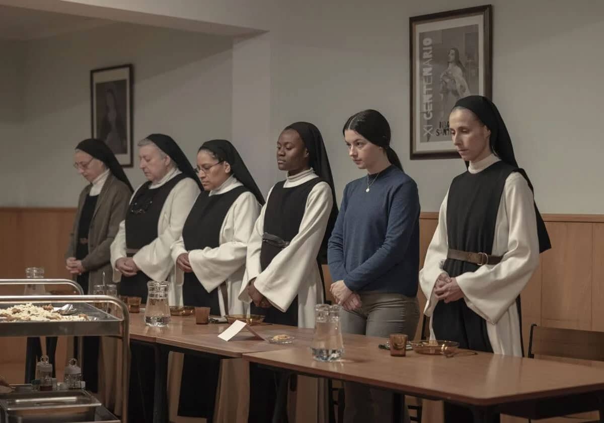 Una fila di sei donne, di cui cinque indossano l'abito religioso da suora, in piedi dietro un lungo tavolo in un refettorio. Sono in posizione di preghiera con le mani giunte e il capo leggermente chino. Sul tavolo sono disposti bicchieri d'acqua e piccoli piatti.